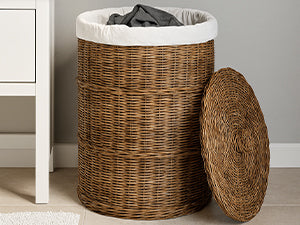 Brauner Rattan-Wäschekorb mit hellem Stoffrand und dunkler Kleidung neben einem weißen Schrank, Deckel rechts